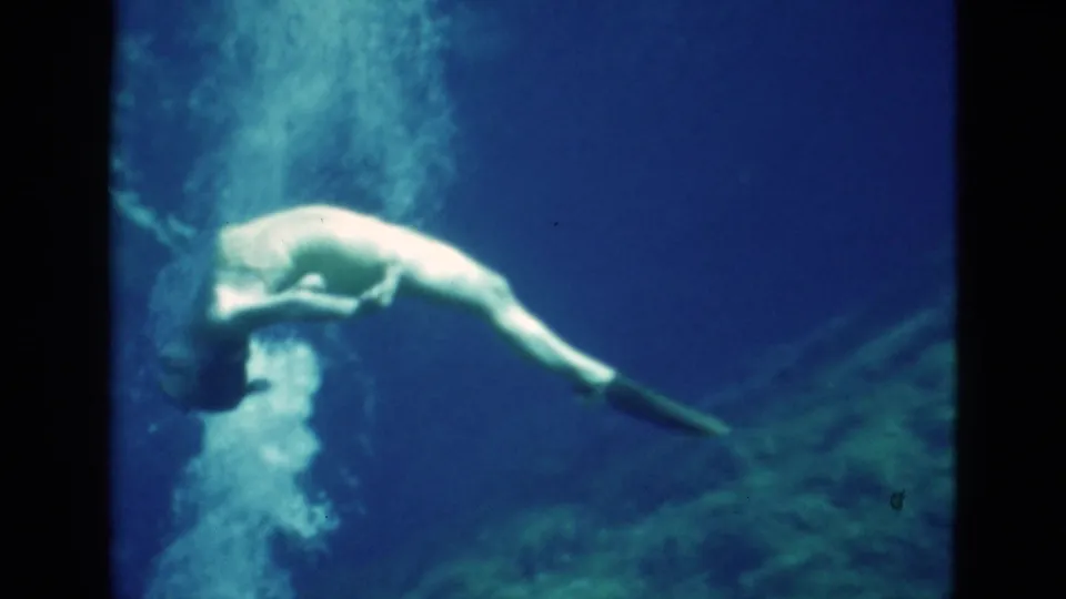 1952 Florida 8mm frame showing a woman underwater curled forward, with swim fins extended and bubbles trailing in blue water.