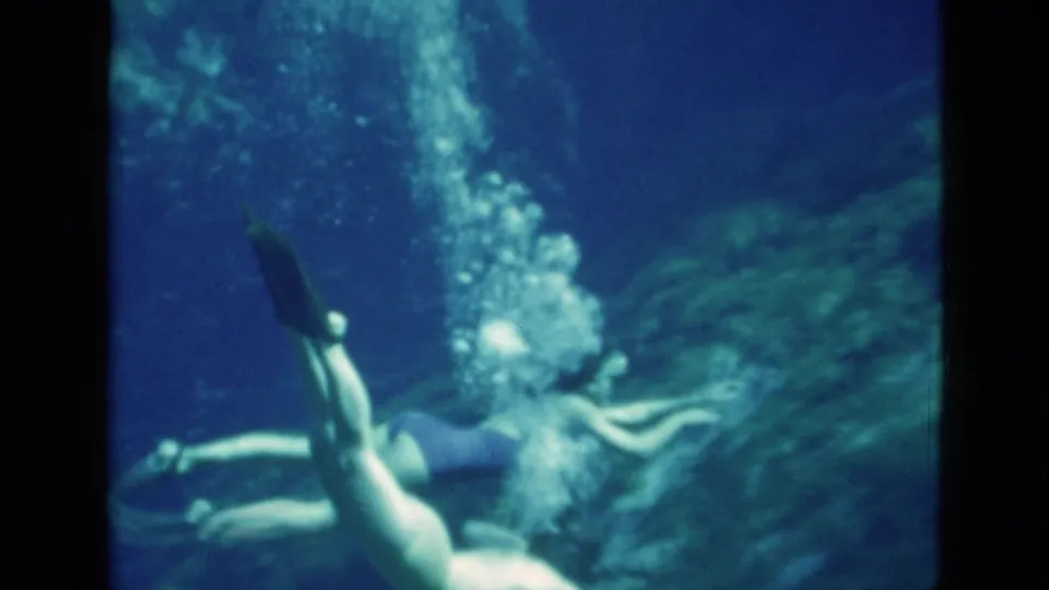 1952 Florida 8mm frame of a woman in mask, dark swimsuit, and fins swimming near a vertical stream of bubbles underwater.