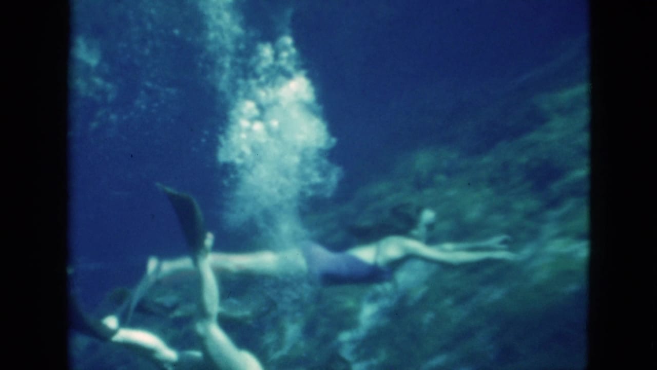 1952 Florida 8mm frame showing a woman in a dark swimsuit swimming underwater with fins, goggles, and bubbles in blue-green water.