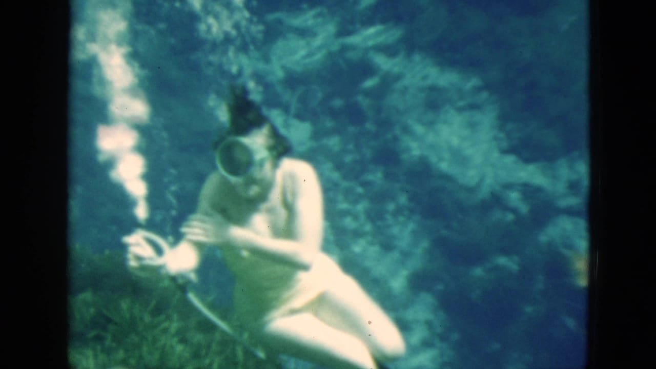 Woman underwater in a diving mask holding a hose near rocks and plants.