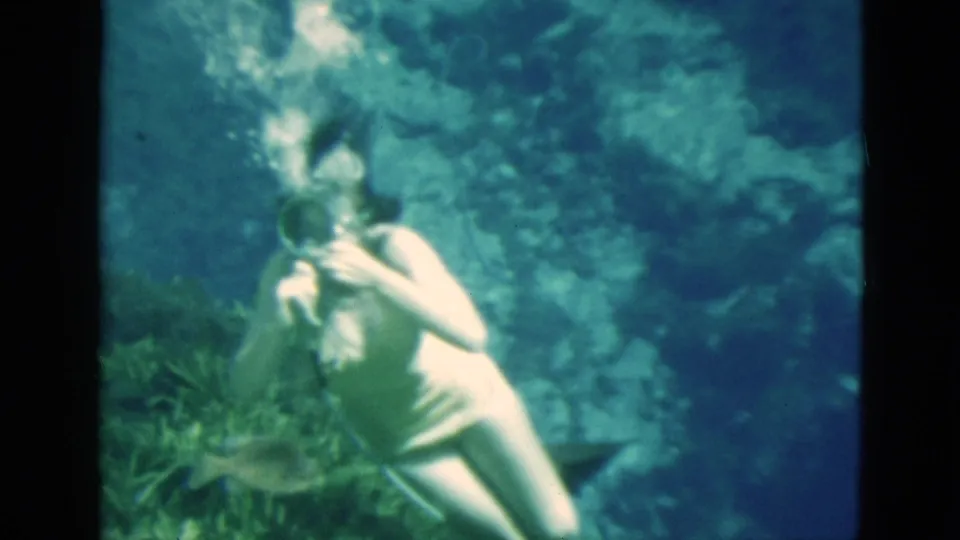 A masked adult woman in Florida 8mm footage holds an air hose underwater, with bubbles rising against a rocky blue-green background.