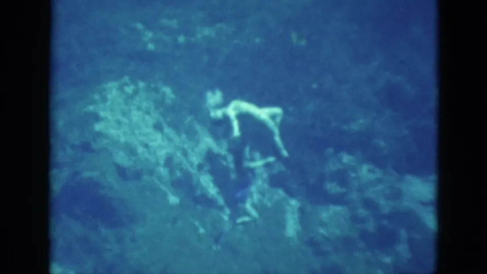 Two swimmers continue an underwater routine near a large rock formation in soft, grainy 1952 Florida home-movie footage.