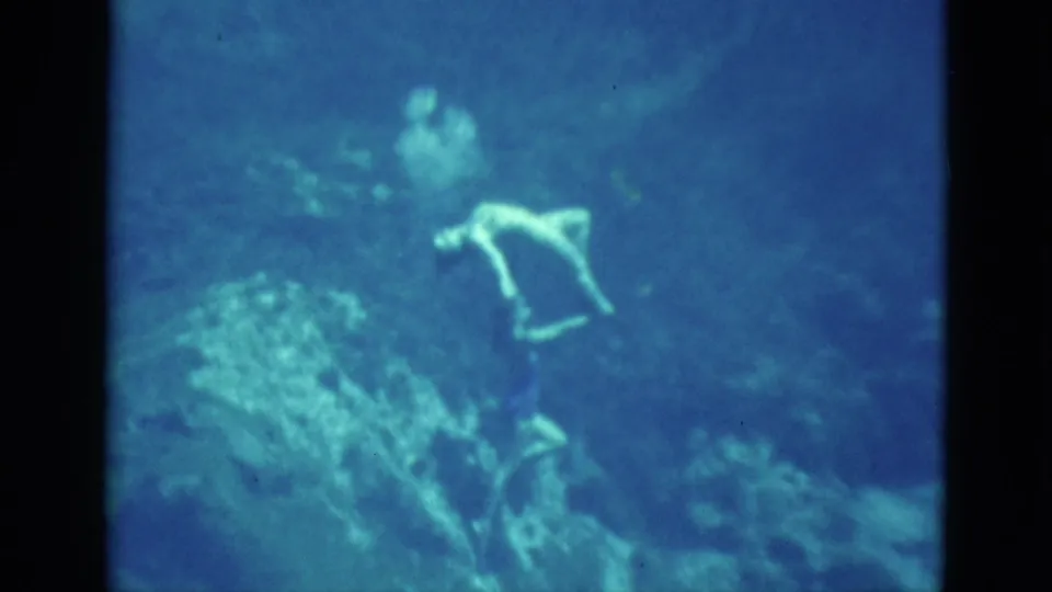 A swimmer bends or turns underwater near rock formations, with another figure faintly visible in the blue-green background.