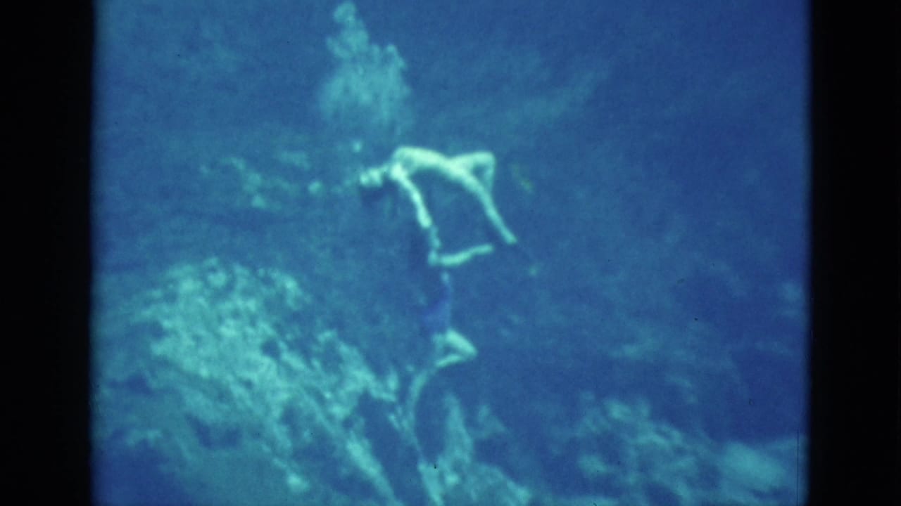 Swimmer underwater in blue water holding an arched pose near pale aquatic shapes.