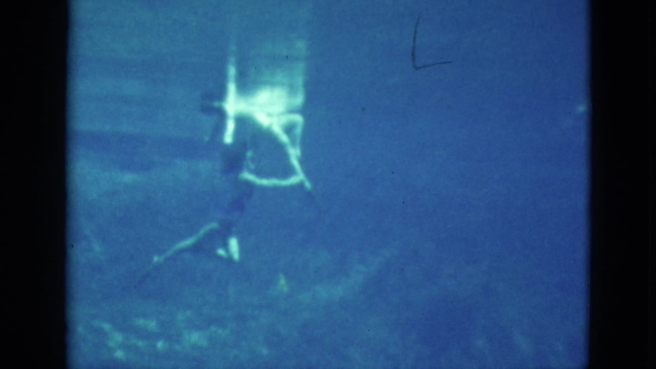 Two young women continue a synchronized underwater movement in the pool, their forms softened by water, grain, and the blue tint of the 8mm scan.