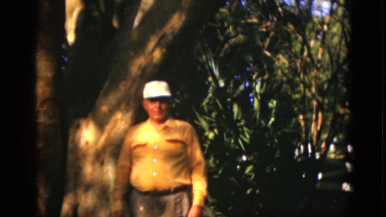 Older man in Quebec home-movie footage standing outdoors near a large tree, wearing a white cap and light yellow shirt.