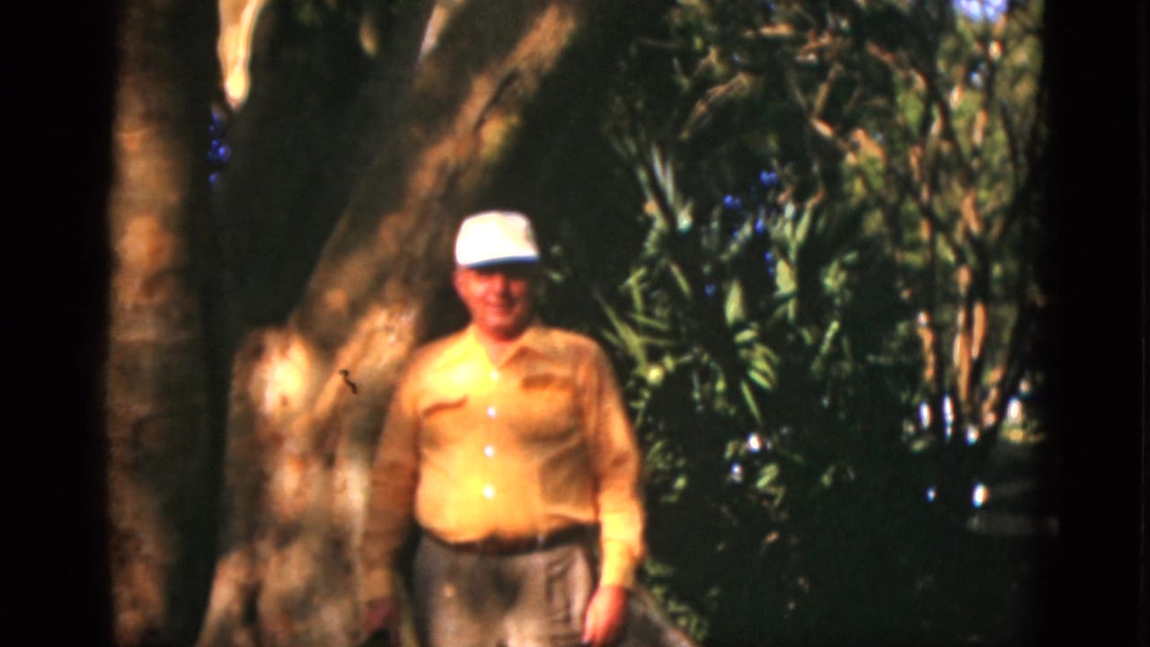Man in a cap standing outdoors beside a large tree trunk.
