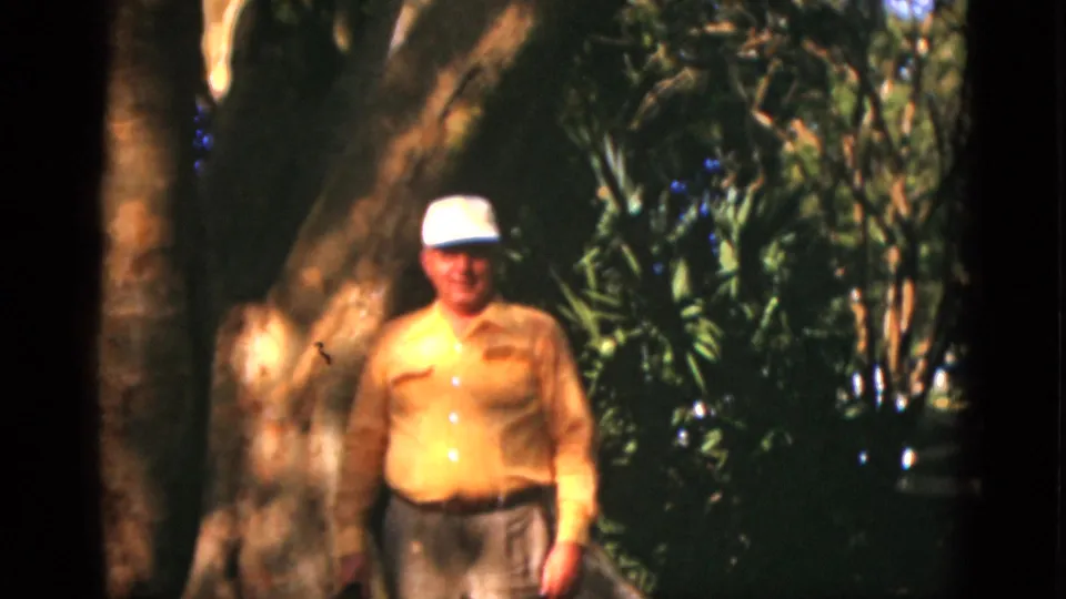 The man remains facing the camera outdoors, framed by a broad tree trunk and shadowed greenery in 1952 Quebec footage.