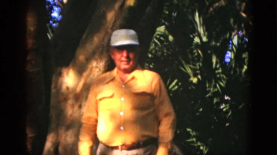 Closer 8mm frame of the older man in a white cap and yellow shirt, standing before a large tree and dark green foliage.