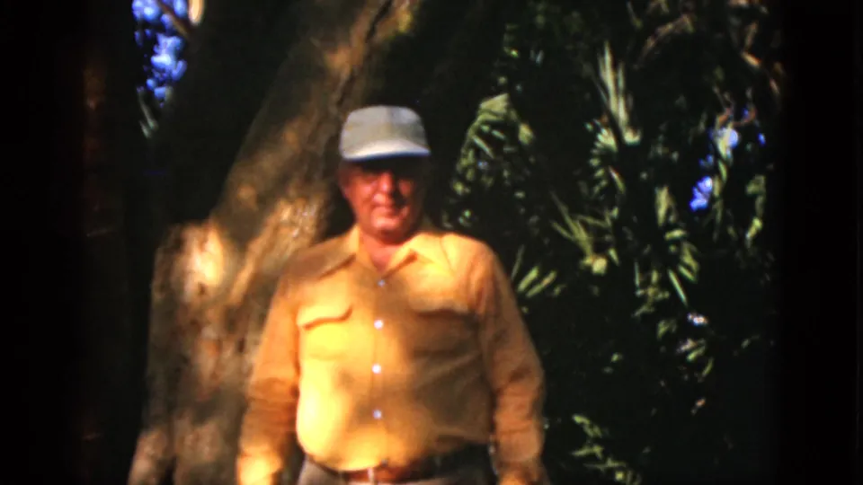 Close outdoor home-movie frame of the older man looking toward the camera, with tree bark and leafy plants behind him.