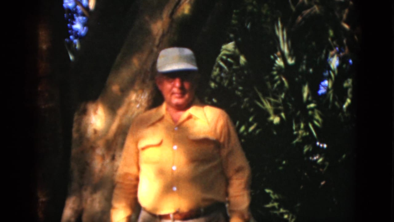 1952 8mm home-movie frame from Quebec metadata showing an older man in a cap standing near a large tree in dense greenery.