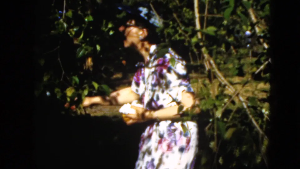 Final 1952 Quebec home-movie frame showing the woman upright among foliage, looking toward branches while holding something light.