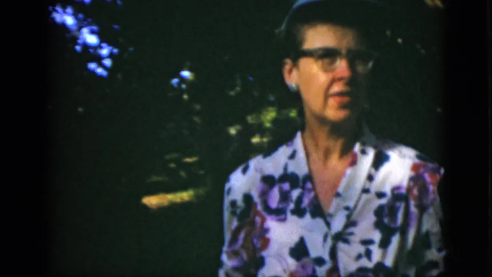 Closer 1952 Quebec home-movie view of the woman in glasses, floral dress, and hat standing in shaded garden light.