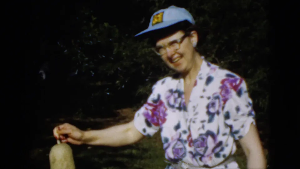 A woman in a floral blouse, eyeglasses, and blue cap lowers the long pod while standing outdoors in the 1952 Quebec home-movie scene.