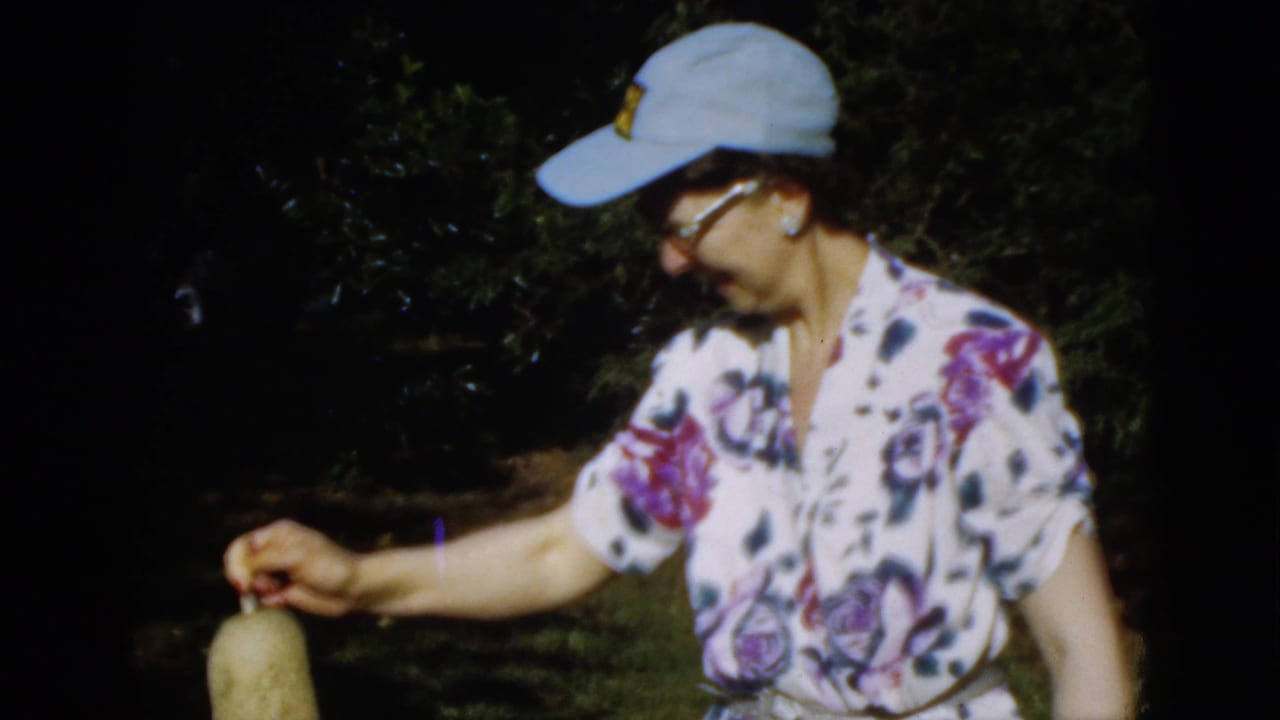 1952 Quebec home-movie frame of a woman in a blue cap and floral blouse handling a plant in a dark, leafy garden.