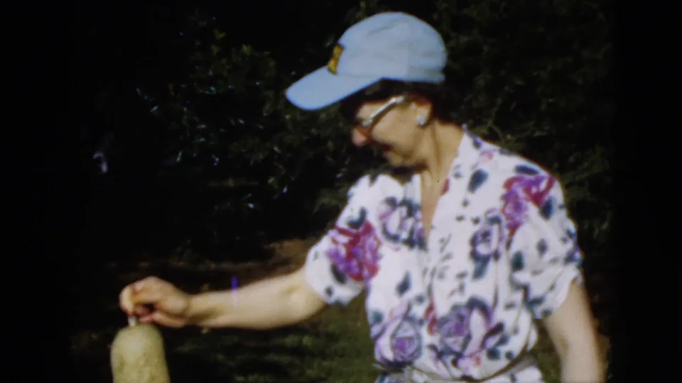 1952 Quebec home-movie frame of a woman in a blue cap and floral blouse handling a plant in a dark, leafy garden.