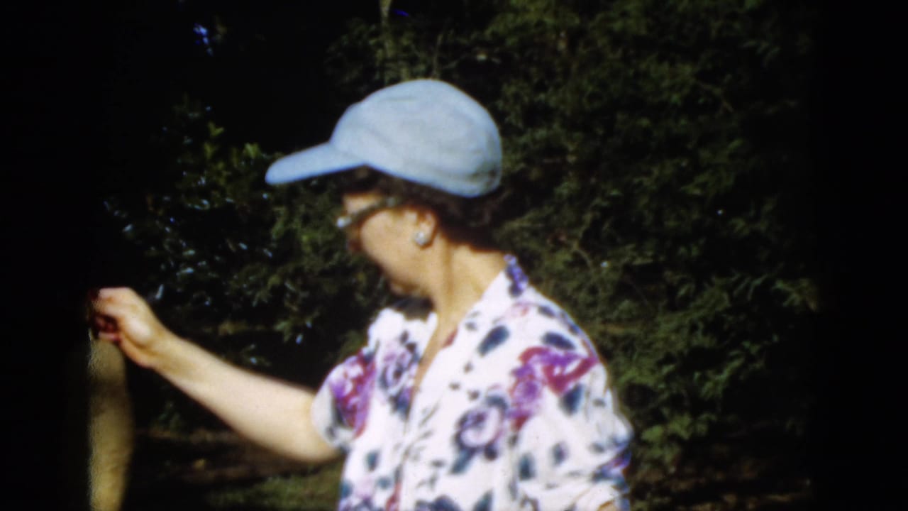 1952 Quebec home-movie frame of a woman in a cap holding a long plant or vegetable in a shaded outdoor garden.