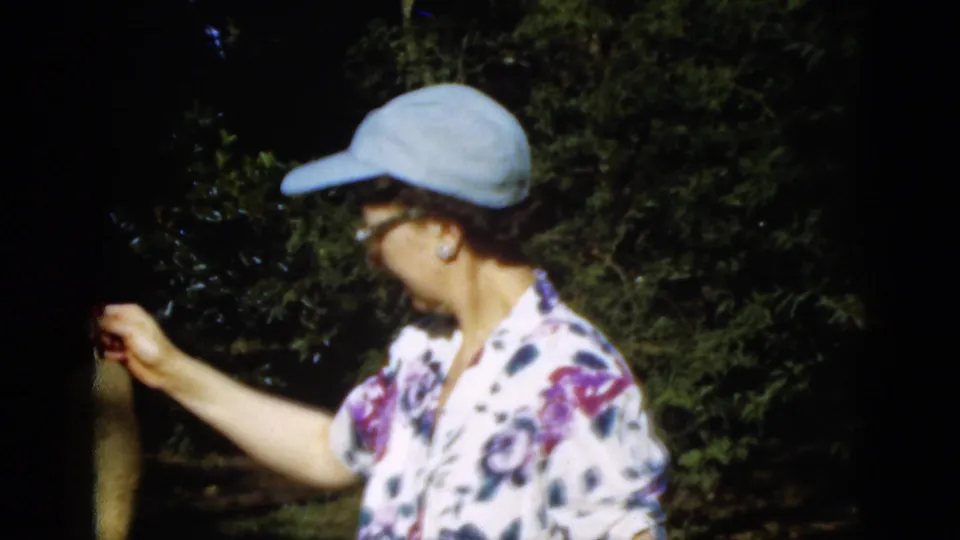 1952 Quebec home-movie frame of a woman in a cap holding a long plant or vegetable in a shaded outdoor garden.