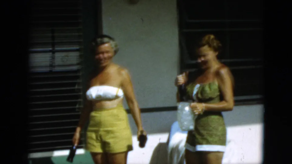 1952 Quebec home-movie frame of two women in retro swimwear standing outdoors near windows and shutters.