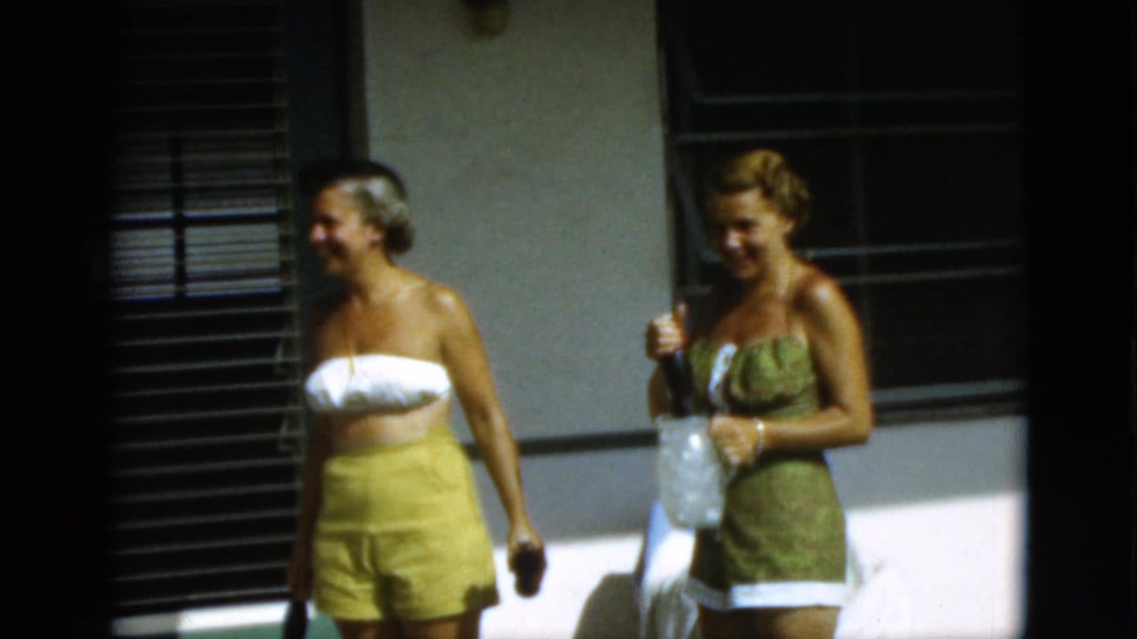 Two women in 1950s swimwear stand on a sunny patio near shaded windows.