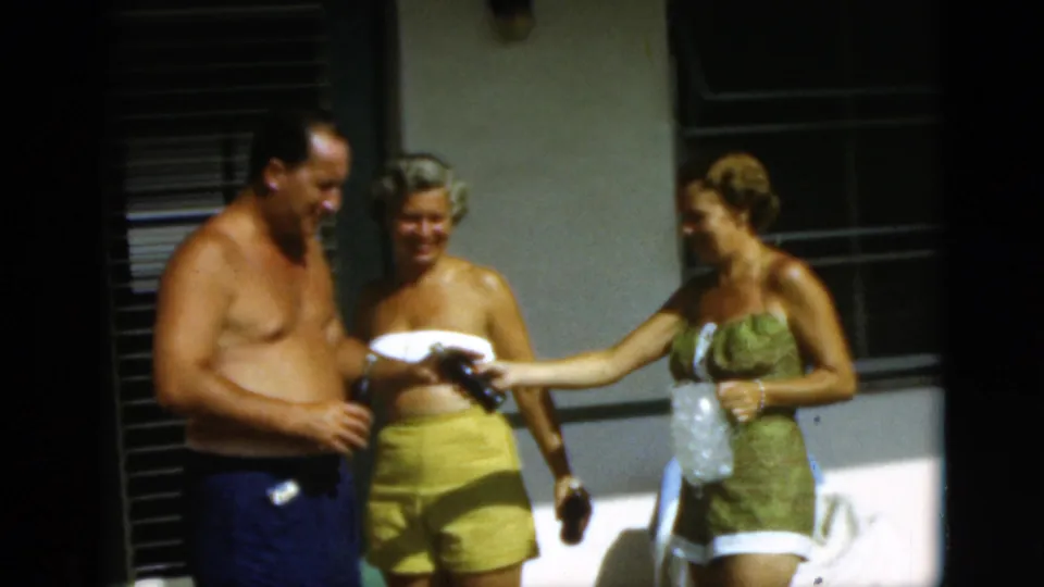 A shirtless man in blue swim trunks stands with the two women while one reaches out with a bottle during the 1952 Quebec patio scene.