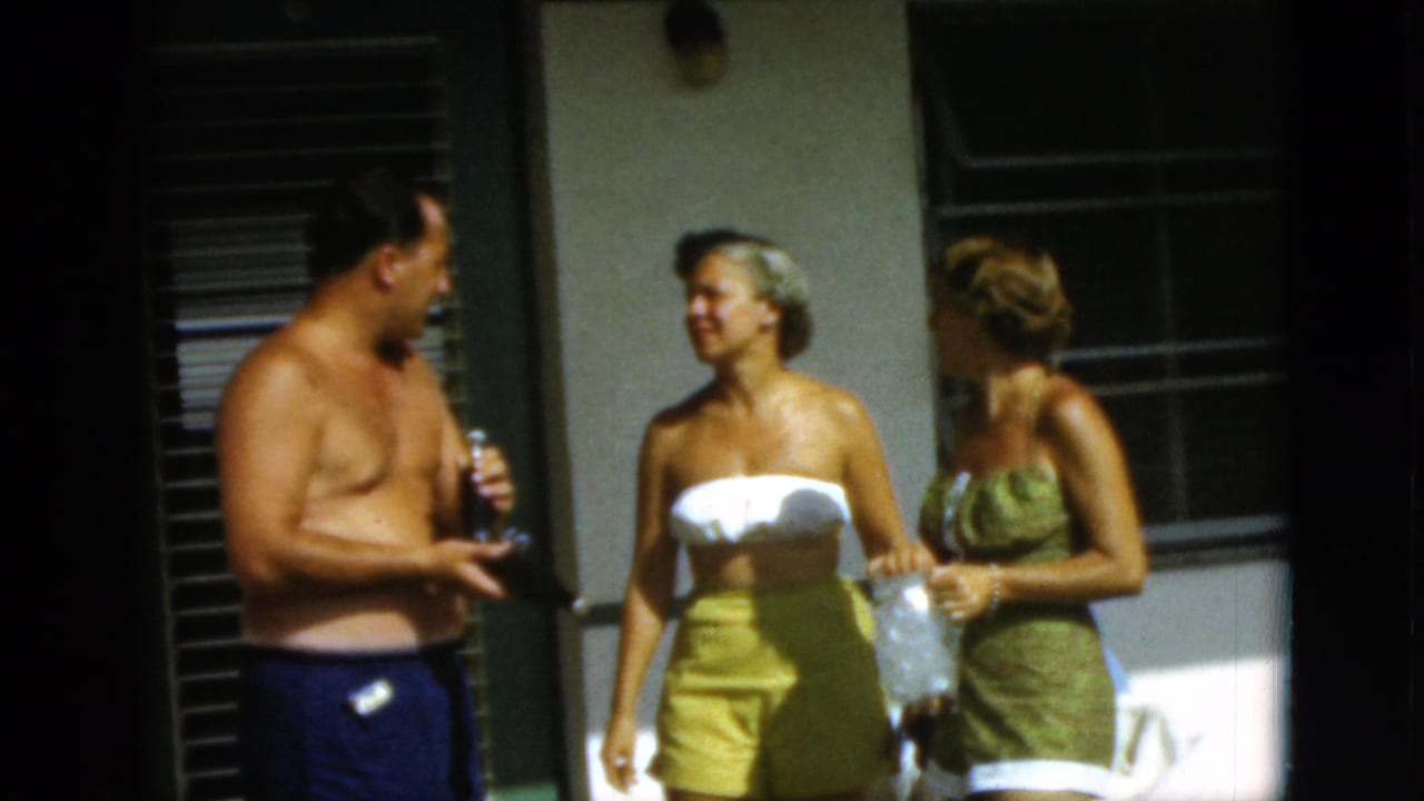 Three adults in swimwear talk outside beside louvered windows in bright sun.