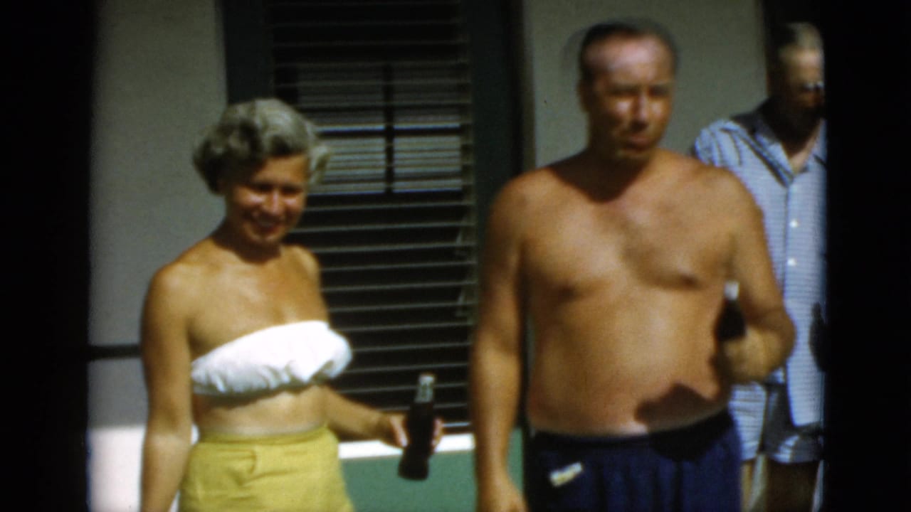 1952 Quebec frame of adults outside, including a woman in a white strapless top and yellow skirt holding a bottle.