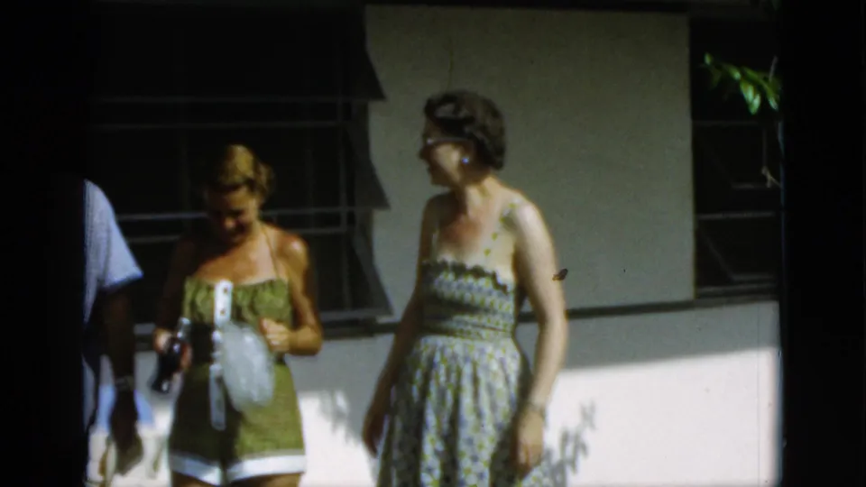 1952 Quebec home-movie frame showing two women in summer dresses beside a patio wall and window.