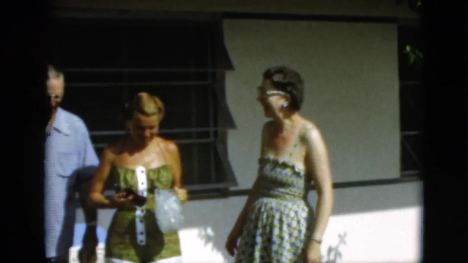 1952 Quebec frame with adults gathered outside, including women in patterned summer clothing near a windowed wall.