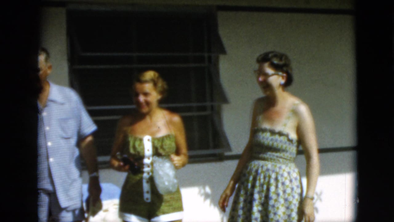 Adults in summer clothing stand and talk outside a building with louvered windows.