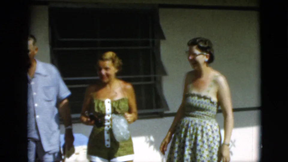 1952 Quebec home-movie frame showing adults in summer clothes conversing near a plain building facade and shaded windows.