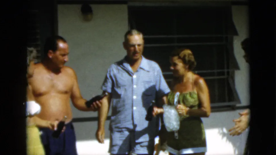 1952 Quebec home-movie frame showing a shirtless man in swim trunks joining adults in conversation outdoors.