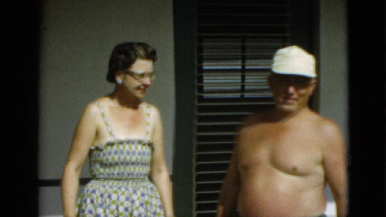 Woman in patterned sundress stands beside shirtless man in white cap near louvered windows.