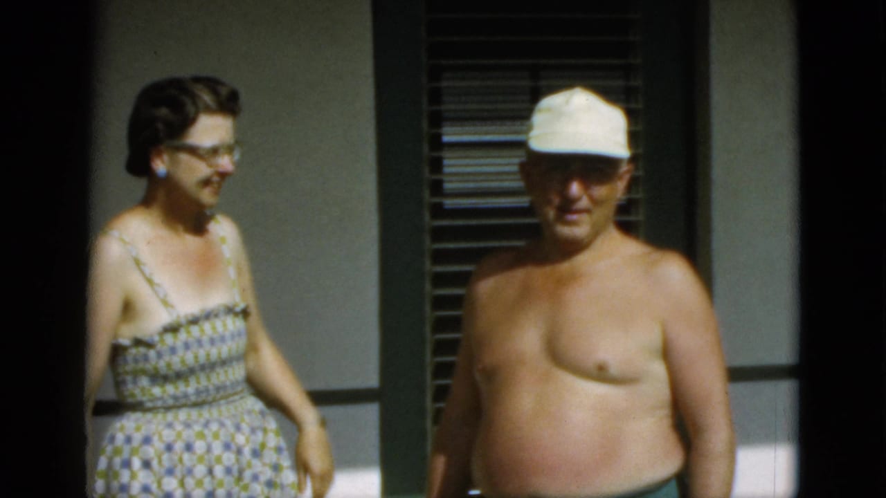 The woman turns toward the shirtless man in a white cap as they stand outside a Quebec building in bright 1952 home-movie footage.