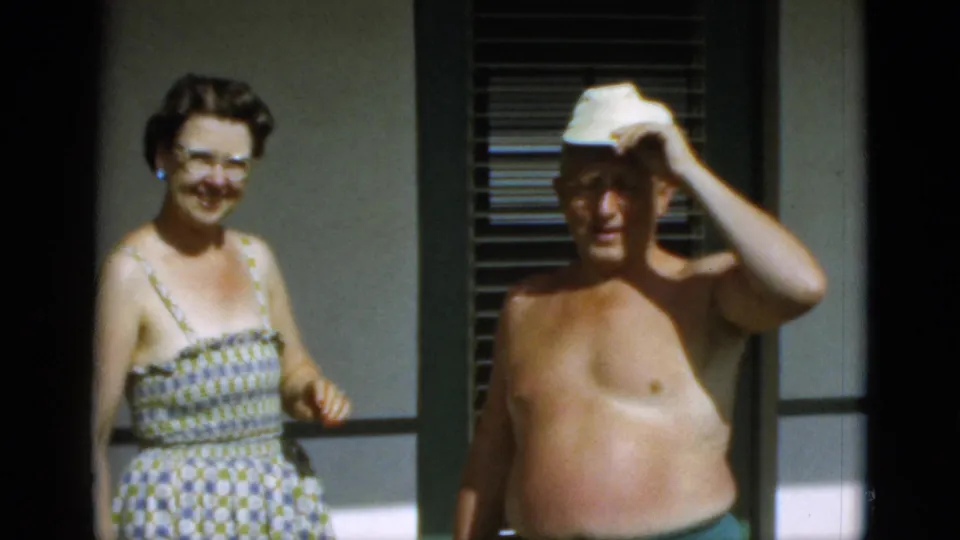 A smiling woman in glasses stands beside a shirtless man adjusting his white cap near louvered windows in 1952 Quebec footage.