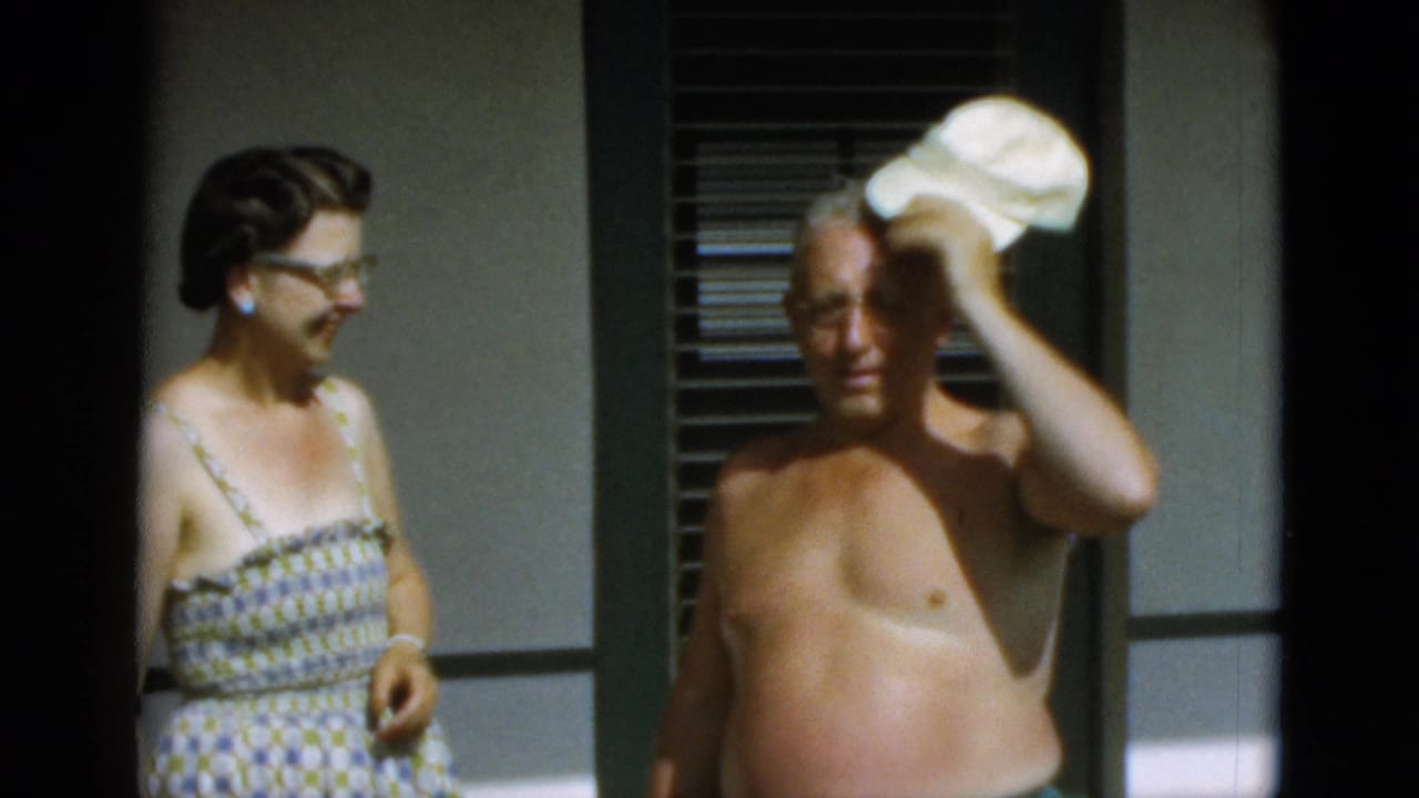 The woman in a patterned dress watches the shirtless man adjust his white cap in bright 1952 Quebec outdoor footage.