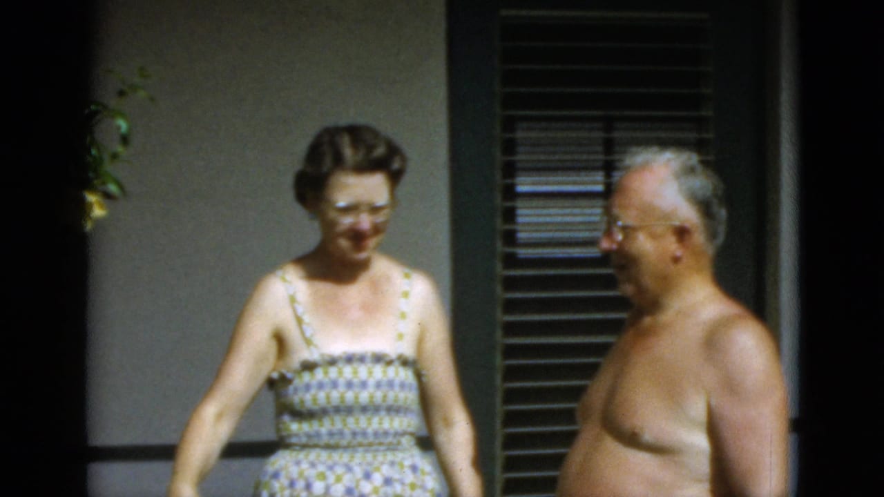 In 1952 Quebec home-movie footage, a woman in glasses and a patterned sundress stands by a shirtless older man outside a wall with blinds.