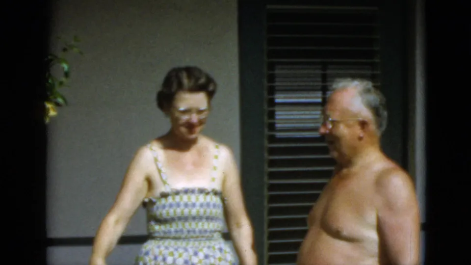 In 1952 Quebec home-movie footage, a woman in glasses and a patterned sundress stands by a shirtless older man outside a wall with blinds.
