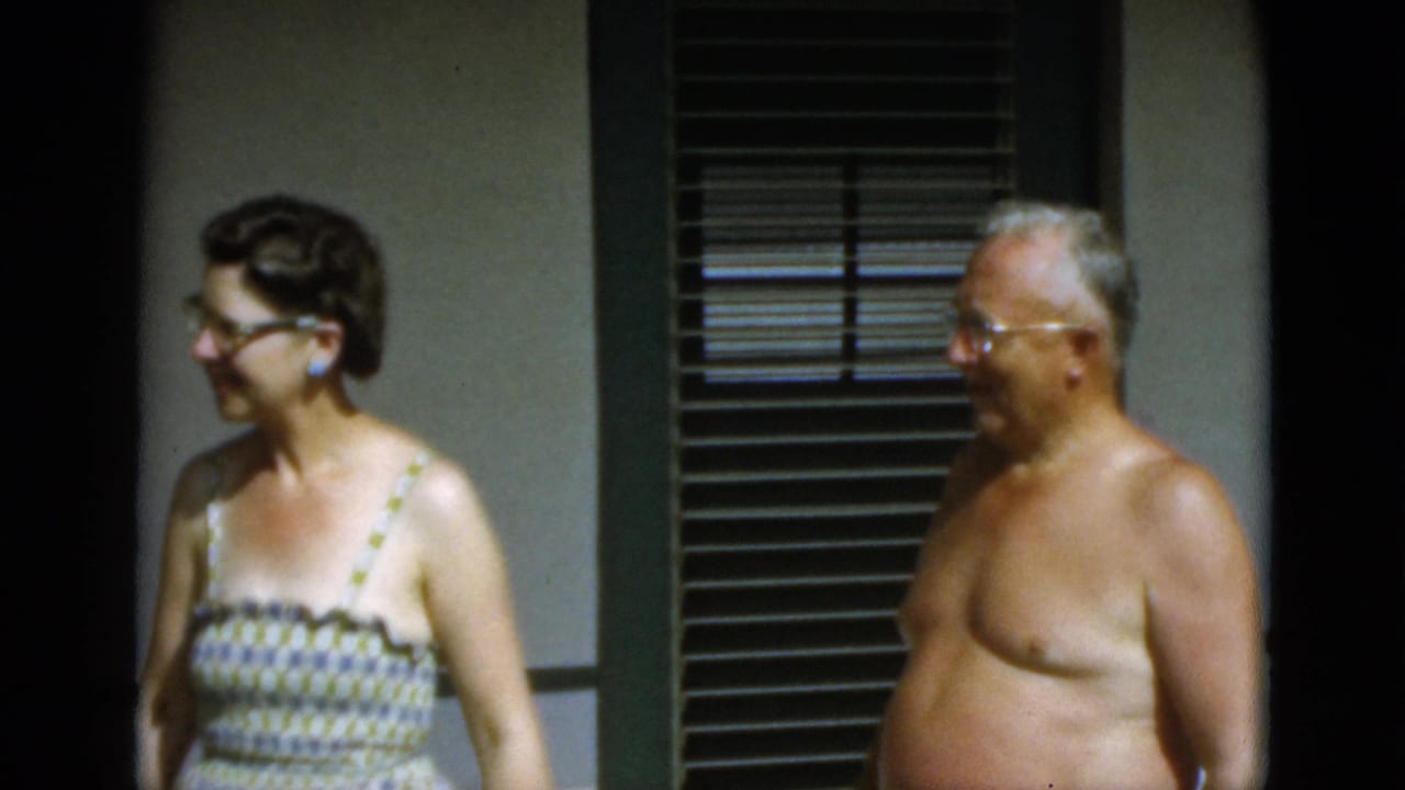 The woman turns and smiles toward the shirtless older man in a brief outdoor domestic moment from 1952 Quebec home-movie footage.