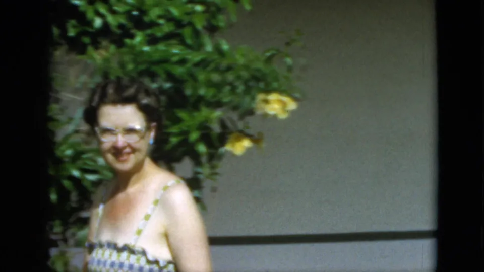 The woman smiles toward the camera beside leafy plants and yellow flowers, continuing the 1952 Quebec domestic home-movie scene.