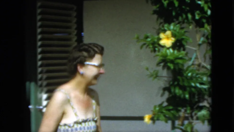 The woman walks across the frame near yellow flowers, exterior wall, and window blinds in the short 1952 Quebec home-movie clip.