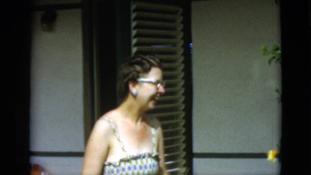 1952 Quebec home-movie frame showing an older woman in glasses and a patterned sundress smiling beside shutters in bright sun.
