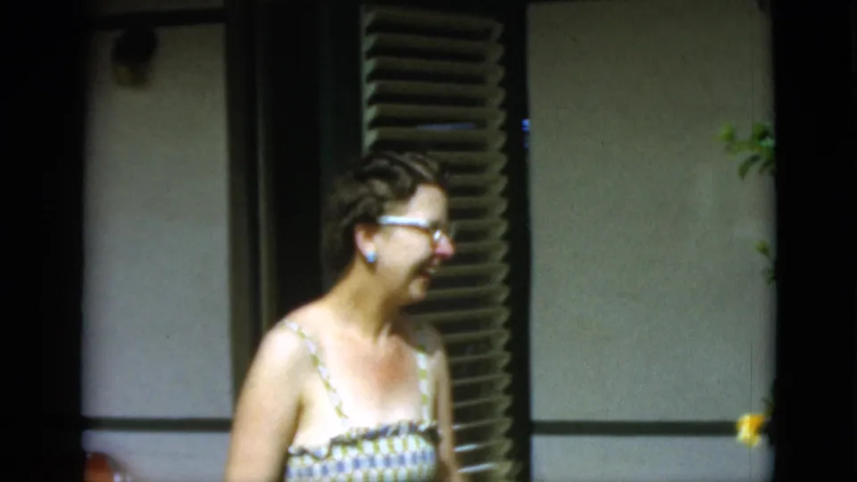 1952 Quebec home-movie frame showing an older woman in glasses and a patterned sundress smiling beside shutters in bright sun.
