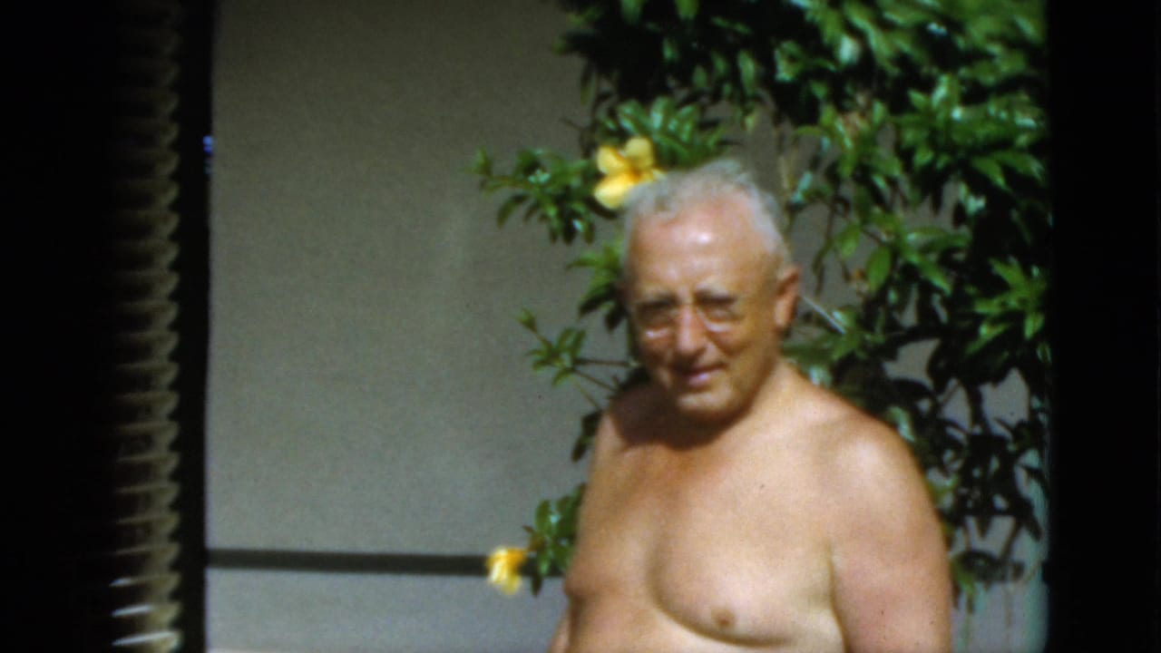 Older shirtless man in glasses stands outdoors beside green foliage and yellow flowers.