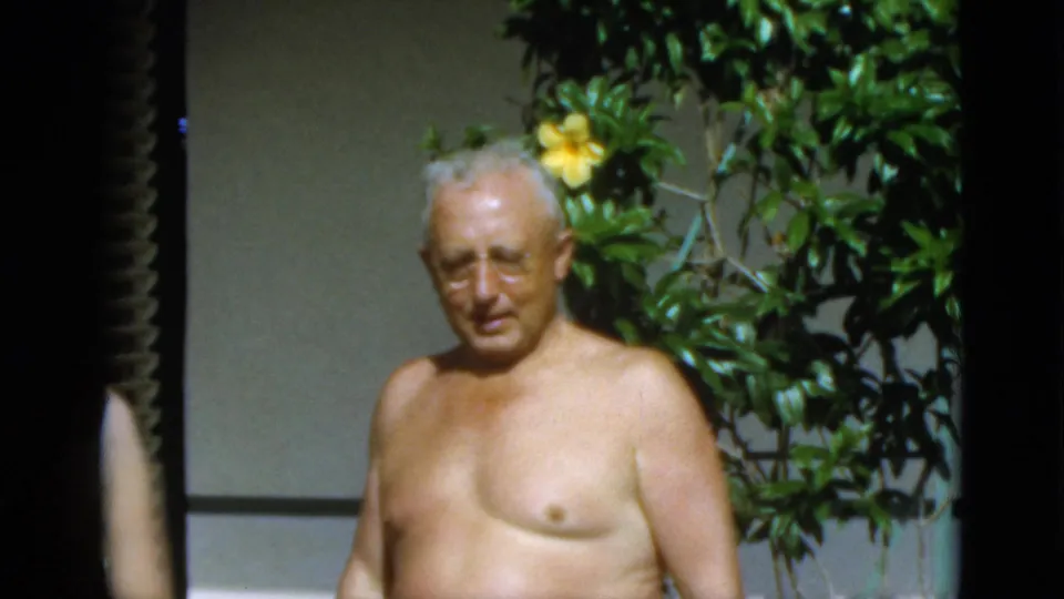 1952 Quebec home-movie frame of an older shirtless man near dense greenery and yellow blossoms, with shutters partly visible.