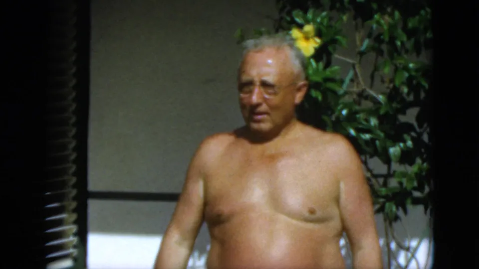 1952 Quebec home-movie frame showing an older shirtless man in glasses outdoors beside shutters and yellow flowers.