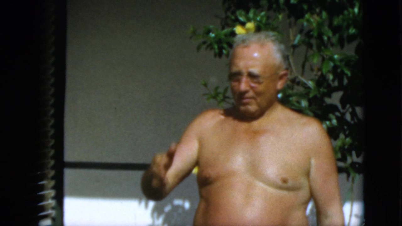 Older shirtless man wearing glasses stands near yellow flowers in bright sunlight.
