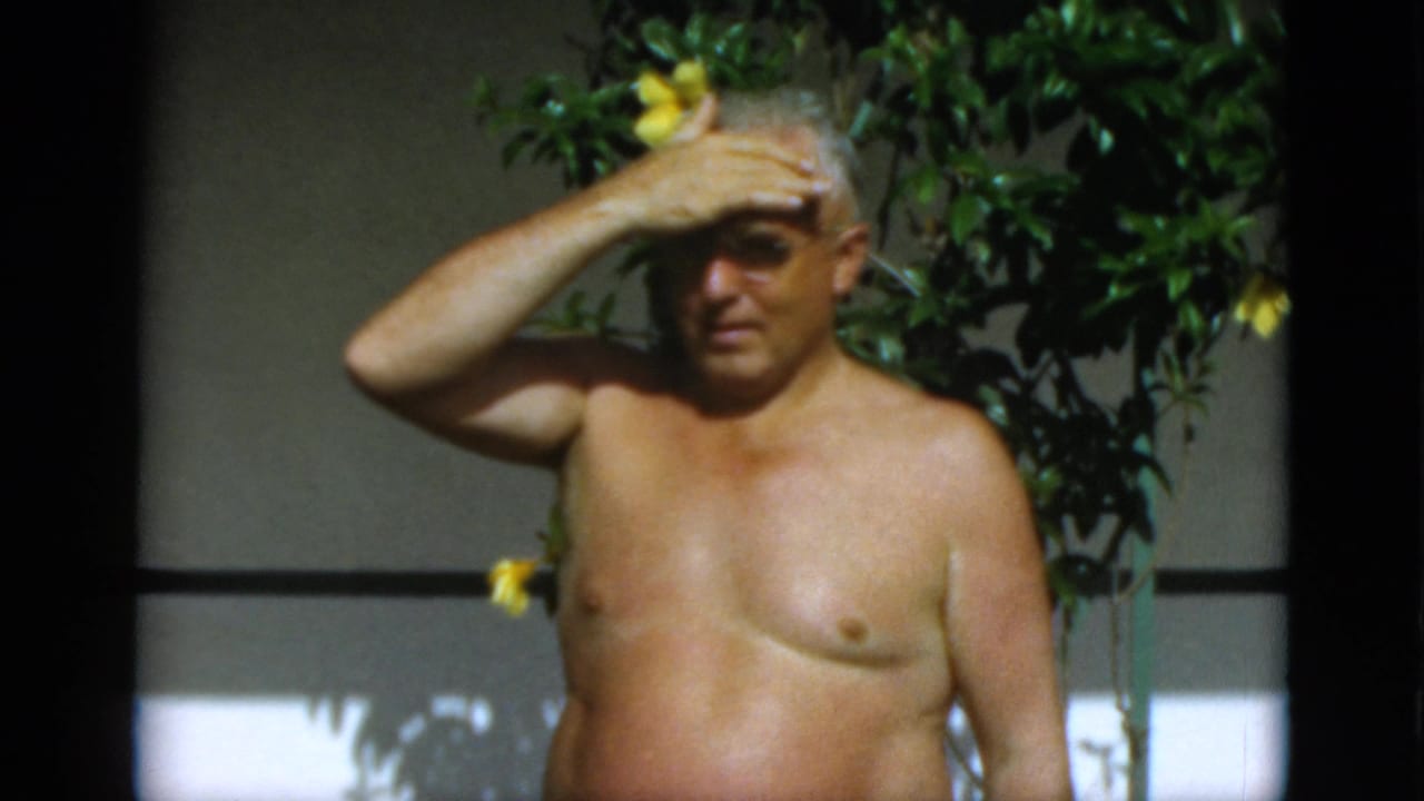 Older shirtless man shades his eyes with one hand beside flowering foliage.