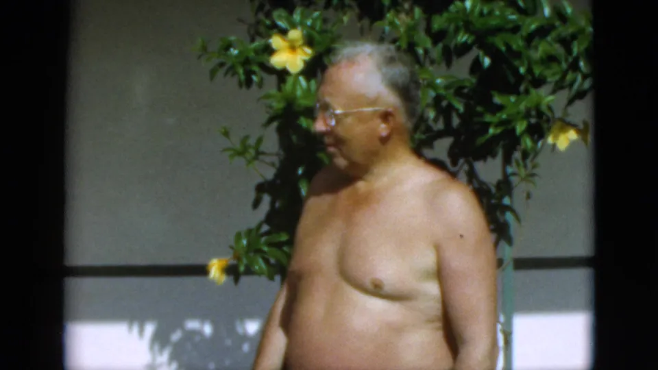 1952 Quebec 8mm frame of an older shirtless man in side view, wearing glasses near a leafy flowering plant.