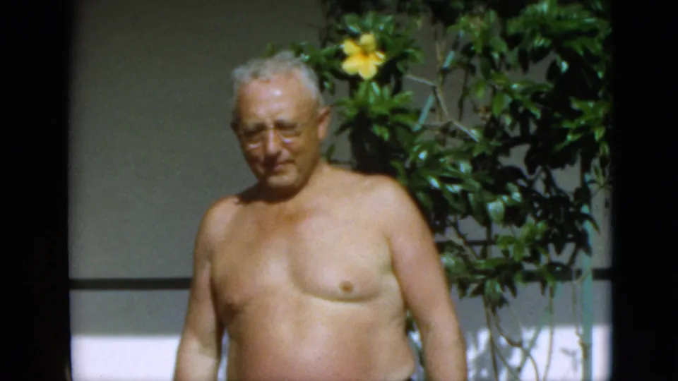1952 Quebec home-movie frame with an older shirtless man facing the camera near sunlit foliage and yellow flowers.